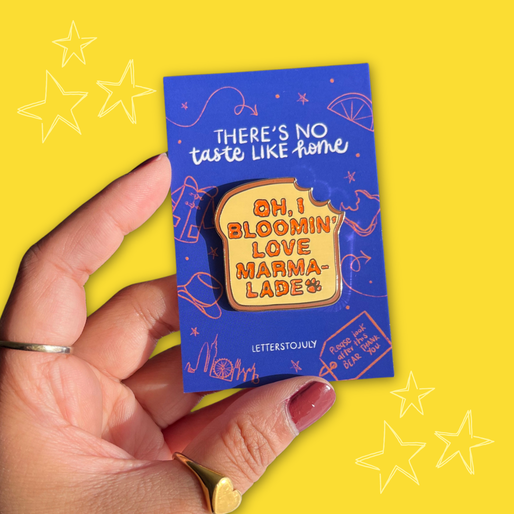 A hand holds a blue enamel pin on its backing card against a bright yellow background. The pin is shaped like a slice of toast with orange lettering that reads, 'OH, I BLOOMIN' LOVE MARMA-LADE'.